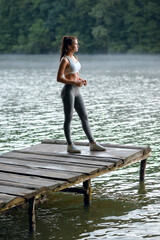 Fototapeta na wymiar Attractive sporty woman in stylish sportswear standing on the wooden pier on the lake and enjoying beautiful nature. Sport concept