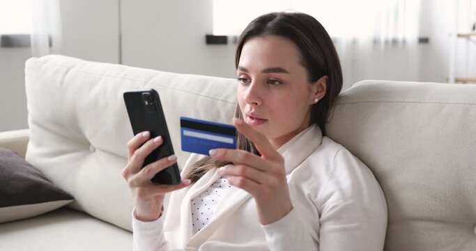 Happy beautiful young woman holding smartphone and plastic banking credit card, entering payment information in mobile application, purchasing goods in internet store or booking hotel, flight tickets.