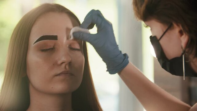 Cosmetologist wipes out the tinted color from the brow of a young woman