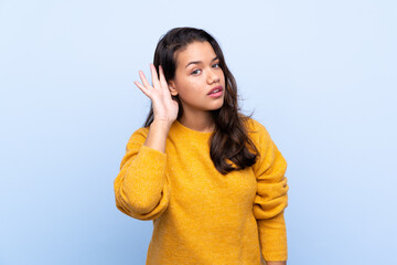 Young Colombian girl with sweater over isolated blue background listening to something by putting hand on the ear