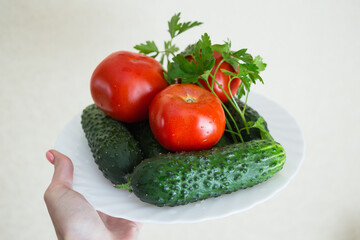 A plate on the woman hand. A fresh tomatoes, sliced cucumbers and sprig of parsley on the white plate on the light table. Seasoning vegetables on a table. Healthy ingredient for salad.