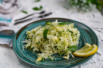 Crispy salad of fresh vegetables with herbs. Young cabbage with cucumber and arugula leaves, decorated with lemon and a bouquet of white flowers on a light textured stone table with Cutlery