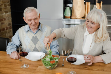 smiling senior couple having dinner with red wine and salad at home on quarantine