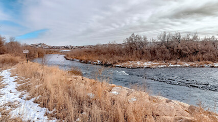 Panorama River with flowing water along grassy and rocky banks dusted with winter snow