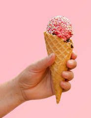 Hand holding strawberry ice cream in waffle cone with candy sprinkles isolated on light pink background, taste of summer concept, frozen dessert, vacation scene
