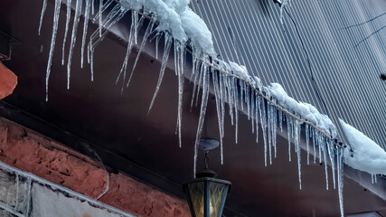 Panorama crop Spiked icicles at the edge of pitched gray roof with clumps of snow in winter