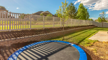 Panorama crop Trampoline at sunlit backyard of home with patio and planting bed against fence