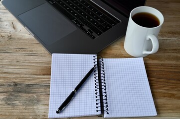 on a wooden table is a laptop with cup of tea and a notebook with pen.