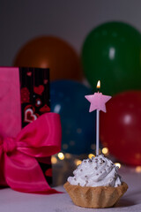festive cupcake with a pink star-shaped candle. On the background of red gift box with a pink ribbon, multi-colored balloons and lights. party, birthday concept. Copy space. . High quality photo