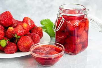 Strawberry jam in a glass jar next to fresh strawberries. On a gray background. Homemade winter fruit blanks. Selective focus.