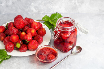 Strawberry jam in a glass jar next to fresh strawberries. On a gray background. Homemade winter fruit blanks. Selective focus.