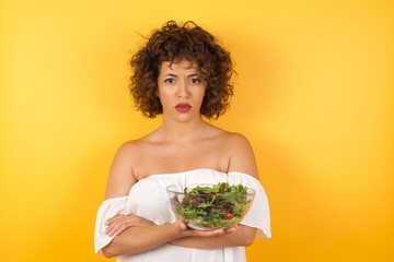 Picture of angry young beautiful woman holding a salad standing isolated over bright background. Looking camera.