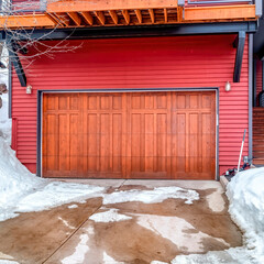 Square Brown garage door and red wall of home with snowy yard and driveway in winter
