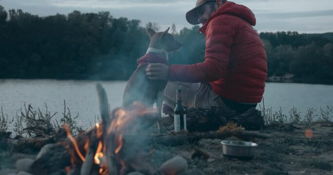Traveling man, urban nomad enjoy beer next to warm campfire during hiking trip.Man and his best friend dog in trendy outdoor outfits watch sunset over lake in forest camping ground. Staycation concept