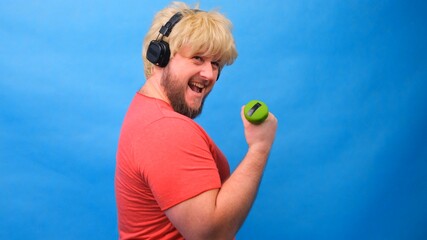 Funny fat man freaky in a wig and a pink t-shirt with dumbbells doing exercises on a blue background.