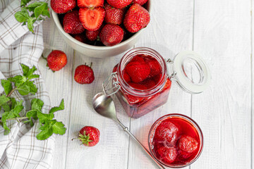 Strawberry jam in a glass jar next to fresh strawberries. On a white wooden background. Homemade winter fruit blanks. Selective focus.
