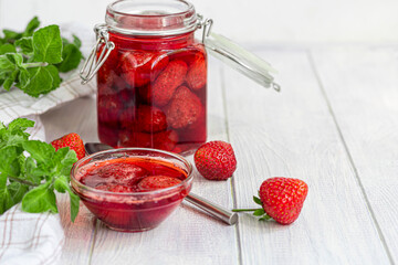 Strawberry jam in a glass jar next to fresh strawberries. On a white wooden background. Homemade winter fruit blanks. Selective focus.