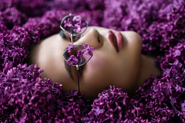 Beautiful model, woman wearing trendy purple sunglasses posing in lilac flowers. Soft focus
