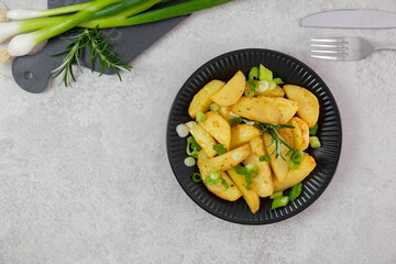 Roasted, baked potatoes on a black plate with fresh green onions and rosemary. Light gray background. Top view. Copy space.