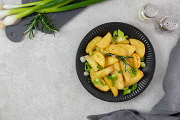 Roasted, baked potatoes on a black plate with fresh green onions and rosemary. Light gray background. Top view. Copy space.