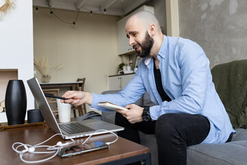 Fototapeta premium A young guy in a blue shirt and black jeans studies online from home using a computer and a diary for notes. Concept of work and study online