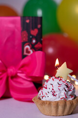 festive cupcake with a yellow star-shaped candle. On the background of red gift box with a pink ribbon, multi-colored balloons and lights. party, birthday concept. Copy space. . High quality photo