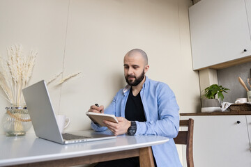 A guy in a blue shirt and black jeans is studying online from home sitting at a desk using a computer and a diary for writing
