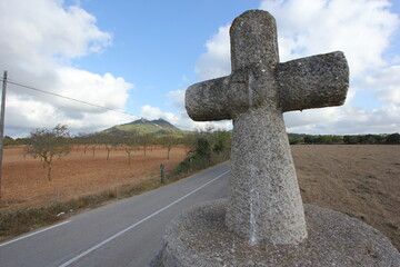 A summer day shot of an old stone cross on the side of a country road with a field with olive trees, a mountain and a blue sky with white clouds in the background. Mallorca island, Spain