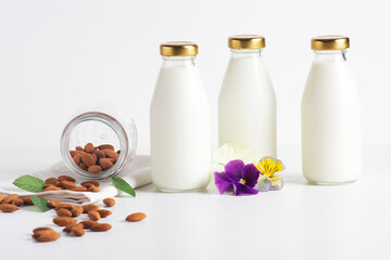 Almond milk in glass bottles stands on a white background, next to it is a jar of almonds and violet flowers. Healthy eating concept, horizontal orientation, close-up.