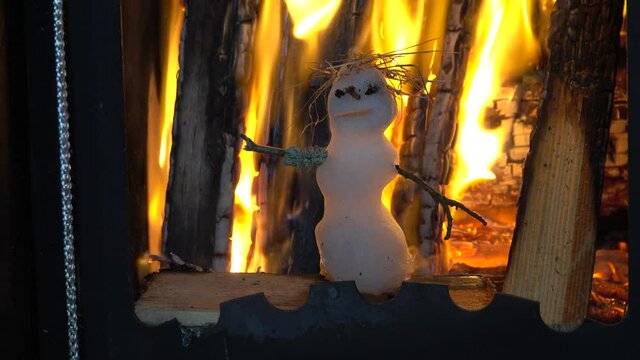 Little snowman melts quickly in hot stove against background of firewood. Concept of victory of heat over cold (spring over winter), heat-loving human (thermophilic person)
