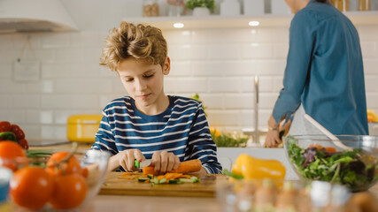 In the Kitchen: Portrait of the Cute Little Boy Cutting Vegetables for the Healthy Dinner. Boy Developing Healthy Habits, eating a lot of Salad and Vegetables. In Background Mom Prepares Dinner