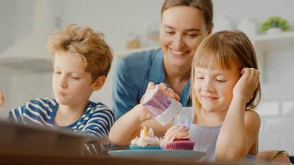 In the Kitchen: Portrait of the Young Mother and Cute Little Daughter Sprinkling Funfetti on Creamy Cupcakes Frosting. Family Cooking Muffins Together. Adorable Children Helping their Caring Parents