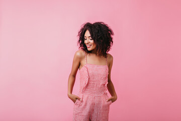 Stylish girl in beautiful striped jumpsuit shyly looks down. Portrait of lady with perfect dark skin on pink background