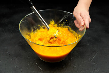 Person cooking pumpkin puree with blender in transparent glass bowl on black table. Mashed pumpkin