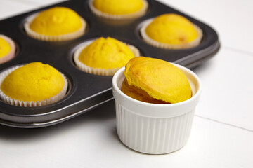 Cupcake in white ceramic ramekin baking bowl. Muffins in metal baking tray on white wooden table