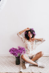 Stunning african girl fooling around during photoshoot with purple flowers. Indoor shot of glamorous black lady in trendy dress sitting on the floor.