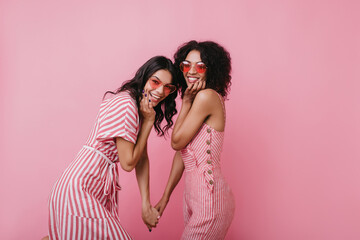 Pretty tanned girls joking during photoshoot on pink background. Indoor photo of refined female models with black hair