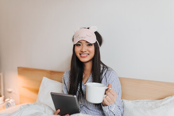 Brown-eyed girl smiles and looks into camera while lying in bed with tablet and cup