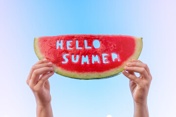 A piece of watermelon against a blue sky. Children's hands are holding a slice of watermelon with...