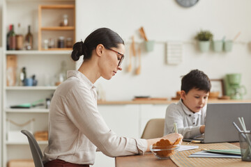 Side view portrait of young mother helping son studying at home while sitting at desk and writing in cozy interior, copy space