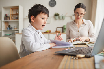 Fototapeta premium Warm-toned side view portrait of cute boy writing in notebook while studying at home in cozy kitchen interior