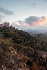 Obraz premium summer afternoon shot of the Puig de Maria sanctuary and monastery standing at the top of the green Sant Marti hills with beautiful mountains and a blue sky with white pink clouds in the back