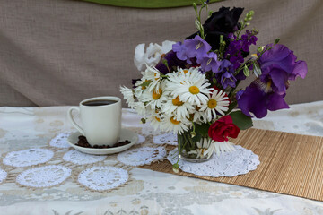 On a white napkin in a glass is a bouquet of daisies, bells and irises. Nearby is a white cup of coffee. On a saucer are coffee beans..