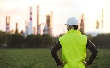 Rear view of young engineer standing outdoors by oil refinery, arms on hips.