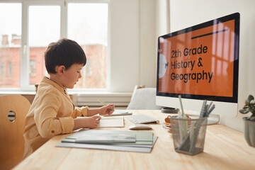 Warm-toned side view portrait of cute little boy studying at home while sitting at desk by computer with online school website, copy space
