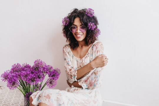 Shy african girl in white dress posing with alliums. Indoor photo of cheerful black young woman in glasses sitting on light background.