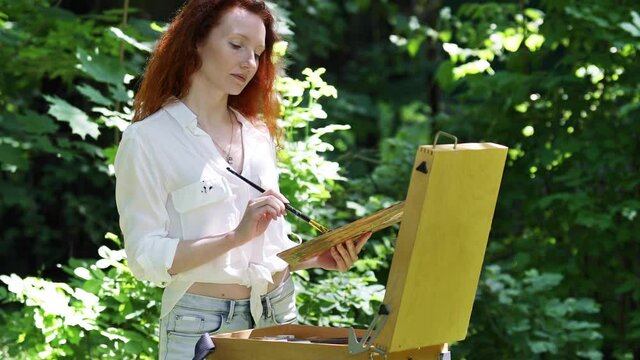 Young red-haired woman draws a picture in the open air in the forest
