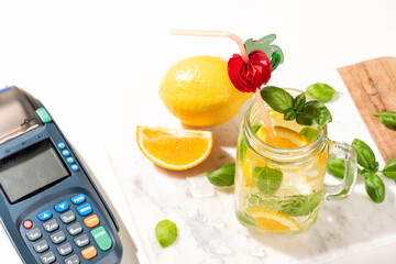 Citrus lemonade or mojito cocktail with lemon and orange and basil and payment terminal. Cold beverage or drink with ice on marble board