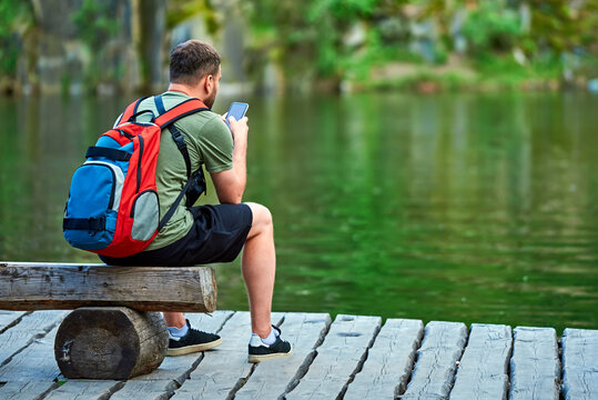 A tourist with a backpack on his back sits on a tree bench by the lake and looks at the phone. beautiful nature and reflection in water. travel photo. Guy in T-shirt, shorts and paints.