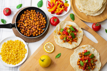 Vegan tortilla tacos with chickpeas, bell peppers, corn, tomatoes, basil leaves and green chilli. On the background are ingredients, lemons, chickpeas in a small pan